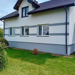 Wojciech Besser Zoom na Dom - Dwukondygnacyjny dom jednorodzinny z jasną elewacją ozdobioną szarymi pasami, ciemnym dachem z dachówki i czarnymi rynnami, otoczony zielonym trawnikiem.