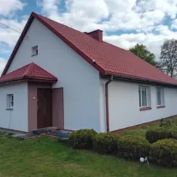 Dom jednorodzinny po remoncie elewacji, z nową, czerwoną dachówką i białymi ścianami. Widoczne elementy wykończeniowe w kolorze brązowym, dopasowane do stolarki okiennej i drzwiowej.