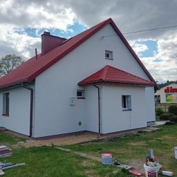 Wojciech Besser Zoom na Dom - Boczny widok odnowionego domu z czerwoną dachówką i białą elewacją. Na trawie widoczne puszki z farbą i narzędzia malarskie, sugerujące świeżo zakończone prace.