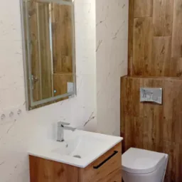 Nowoczesna łazienka z drewnopodobnymi płytkami i podwieszaną toaletą, umywalka z szafką z drewnianym fornirem, lustro z podświetleniem LED.