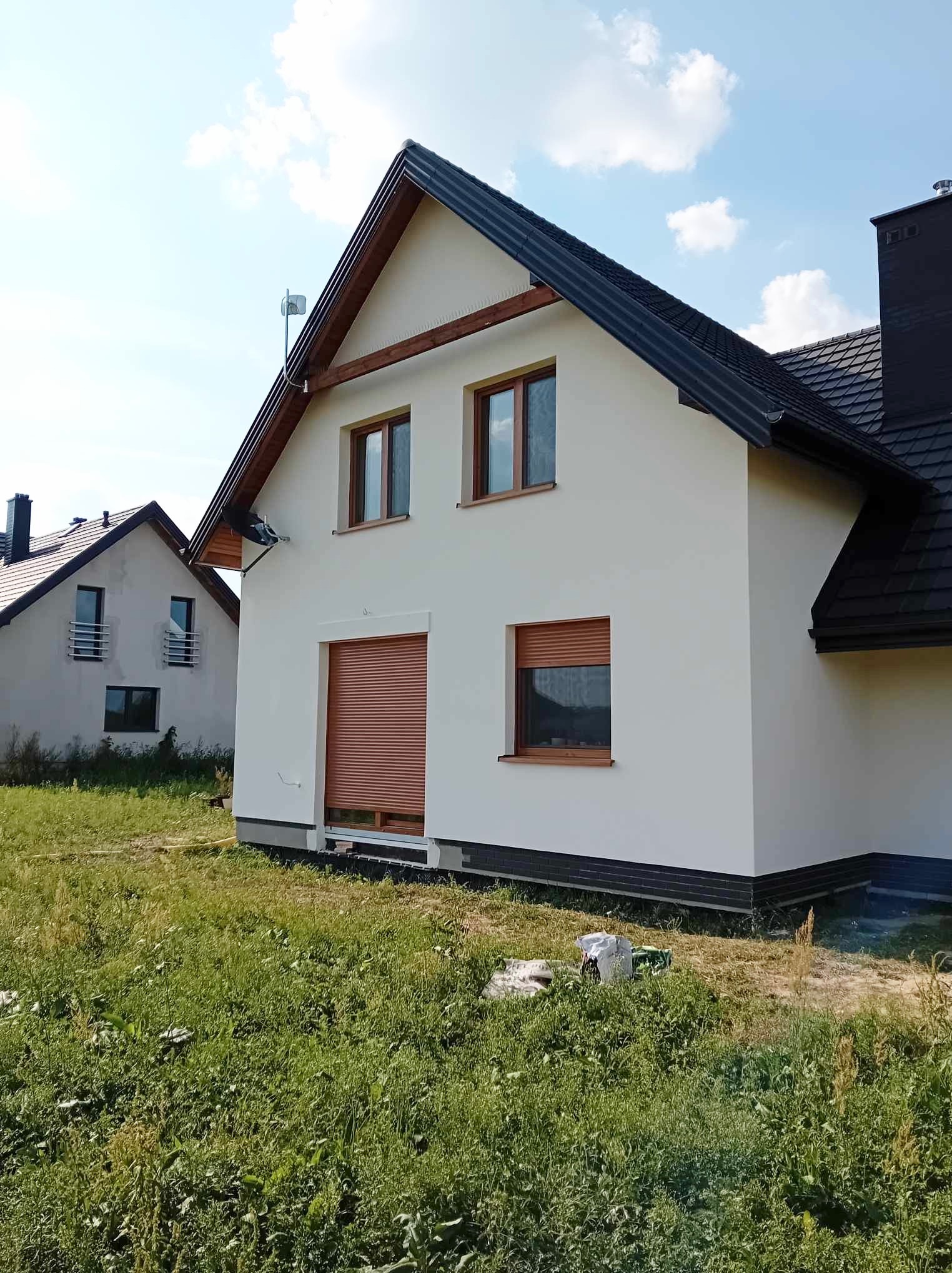 Nowoczesny dom jednorodzinny z jasną elewacją i brązowymi roletami zewnętrznymi, widok z poziomu trawnika, drugi dom w tle.