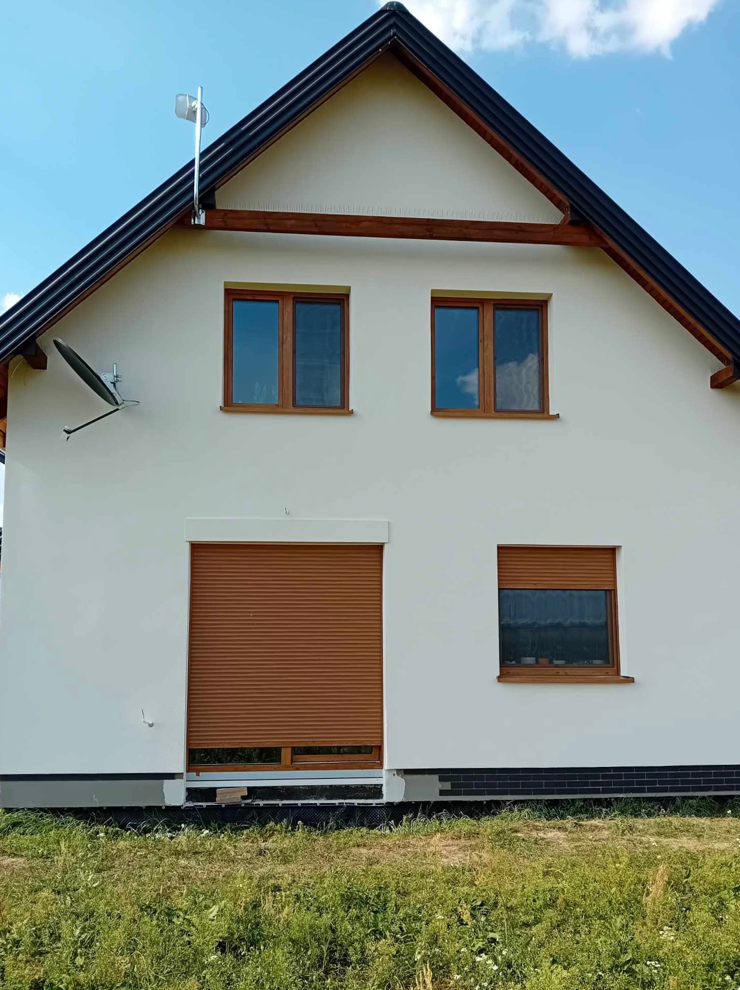 Dom jednorodzinny z białą elewacją, brązowymi oknami i roletami, na zewnątrz widoczny trawnik, antena satelitarna i oświetlenie zewnętrzne.