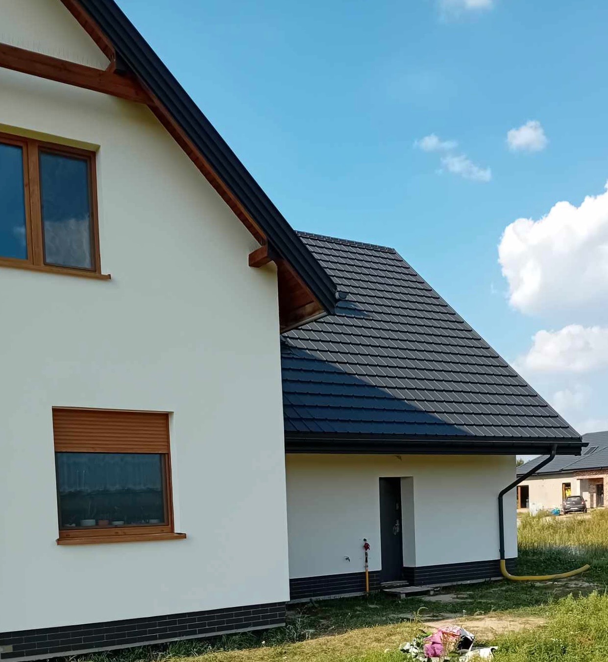 Nowoczesny dom jednorodzinny z białymi ścianami i grafitowym dachem, widok z boku na elewację boczną z oknami z roletami i fragmentem ogrodu.