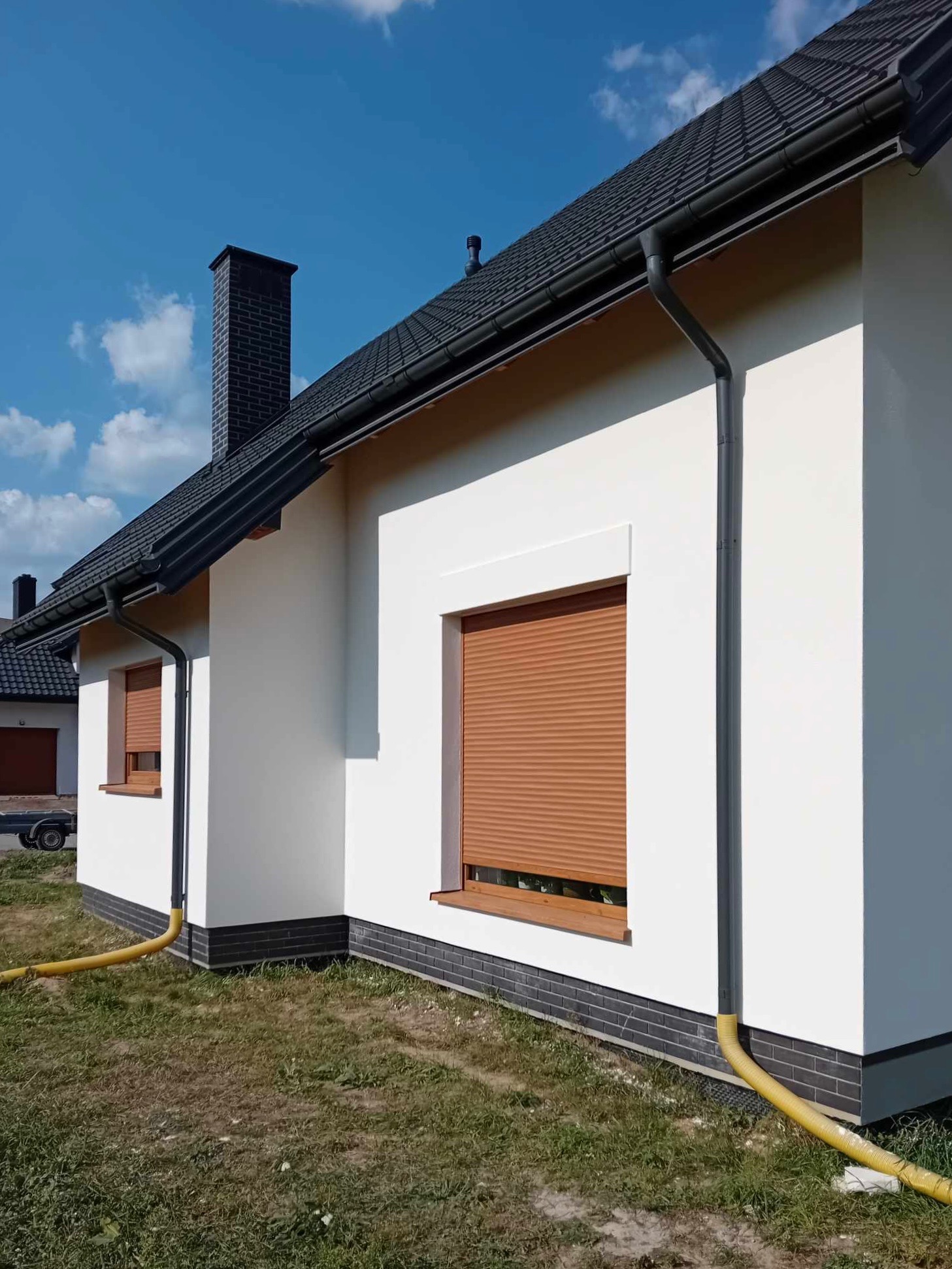 Świeżo pomalowana, biała elewacja domu jednorodzinnego z brązowymi roletami zewnętrznymi i czarnymi elementami dachu oraz ceglaną podmurówką, widok z boku, wąż odprowadzający wodę leży na trawie.