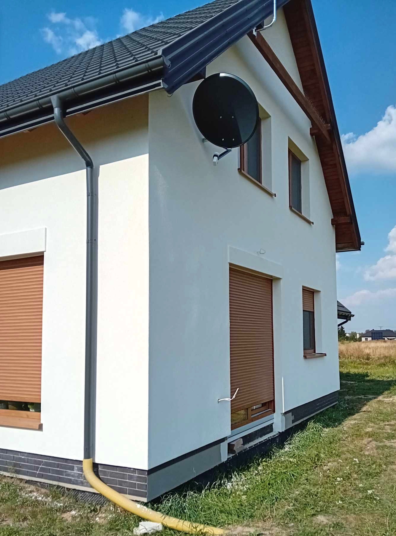 Świeżo pomalowana, biała elewacja domu jednorodzinnego z brązowymi roletami i anteną satelitarną na ścianie, widok z zewnątrz w słoneczny dzień, z widocznym fragmentem dachu i rynny.