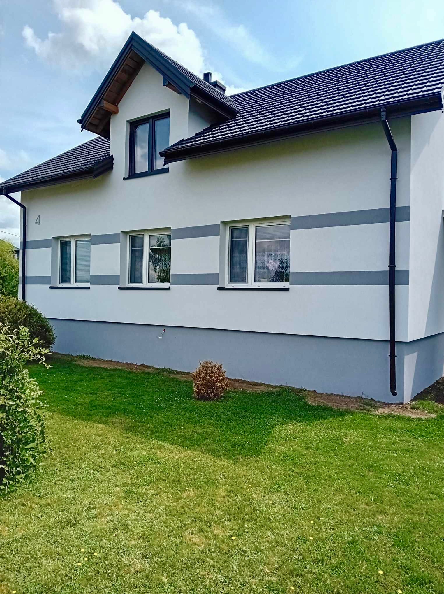 Dwukondygnacyjny dom jednorodzinny z jasną elewacją ozdobioną szarymi pasami, ciemnym dachem z dachówki i czarnymi rynnami, otoczony zielonym trawnikiem.