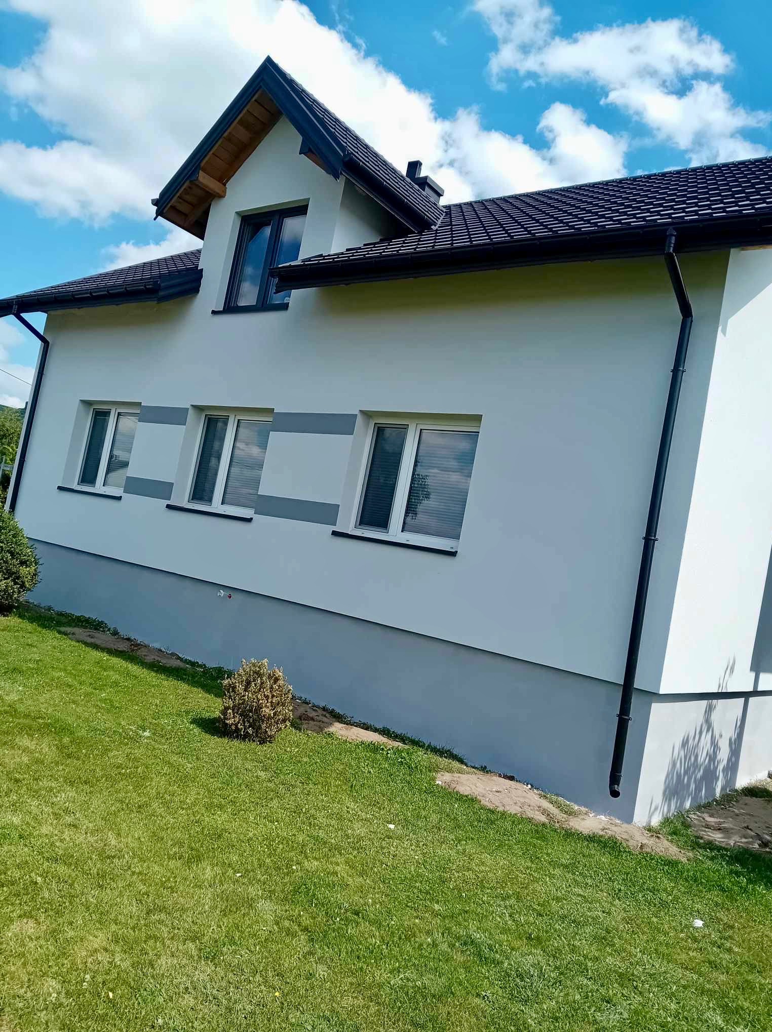 Świeżo pomalowana elewacja domu jednorodzinnego w kolorze białym i szarym, z widocznymi trzema oknami i czarną rynną na tle zielonego trawnika i błękitnego nieba.