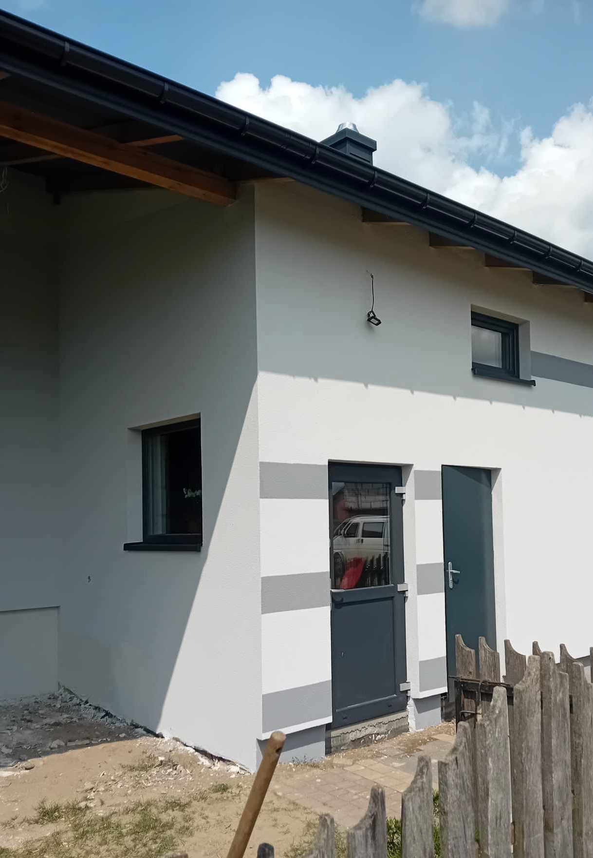 Świeżo pomalowana elewacja budynku z szarymi pasami, widoczne dwie pary drzwi w kolorze antracytowym i okno z czarną ramą, teren wokół budynku częściowo uporządkowany, drewniane ogrodzenie...