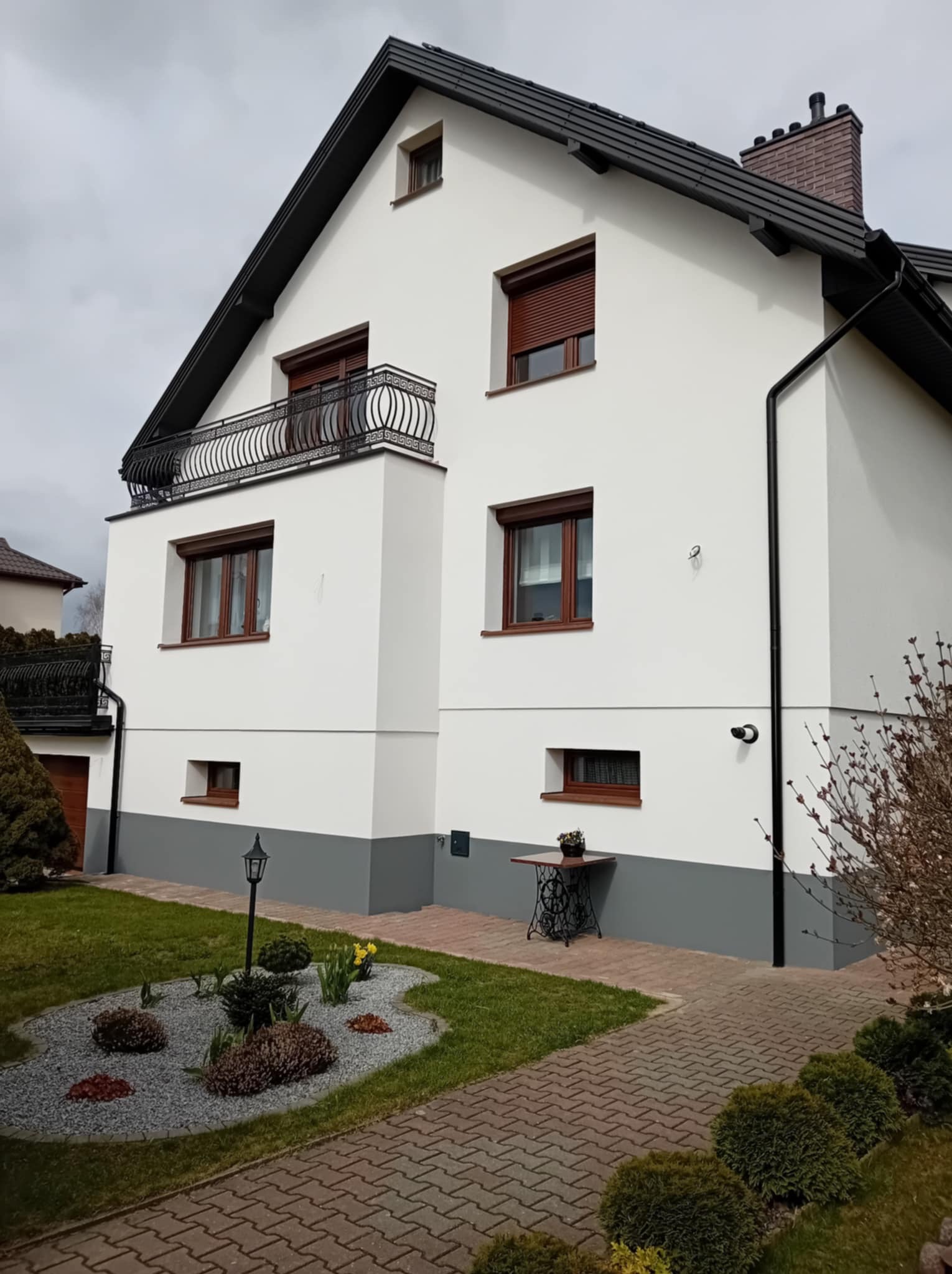 Biała elewacja domu z ciemną podmurówką i dachem, balkon z ozdobną balustradą, brązowe okna z roletami, zadbany ogród z lampą i stolikiem. Widok z poziomu gruntu.