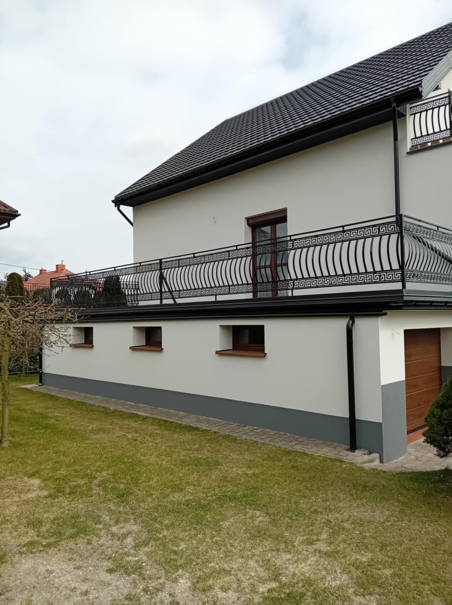 Biały dom z balkonem ozdobionym czarną, dekoracyjną balustradą z motywem meandrycznym. Widoczne okna piwniczne i garaż. Trawa na pierwszym planie.