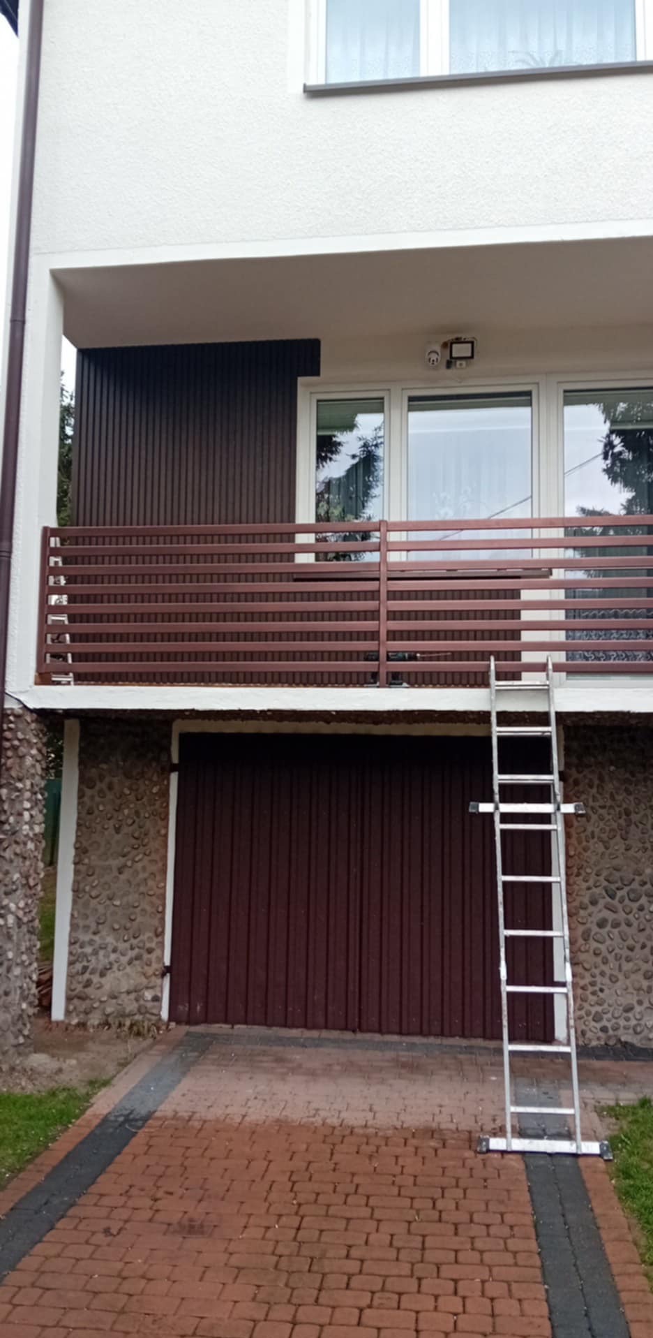 Dwupiętrowy dom z brązowymi elementami elewacji i balkonem. Aluminiowa drabina oparta o ścianę. Widoczne okna z refleksami. Dom po renowacji.