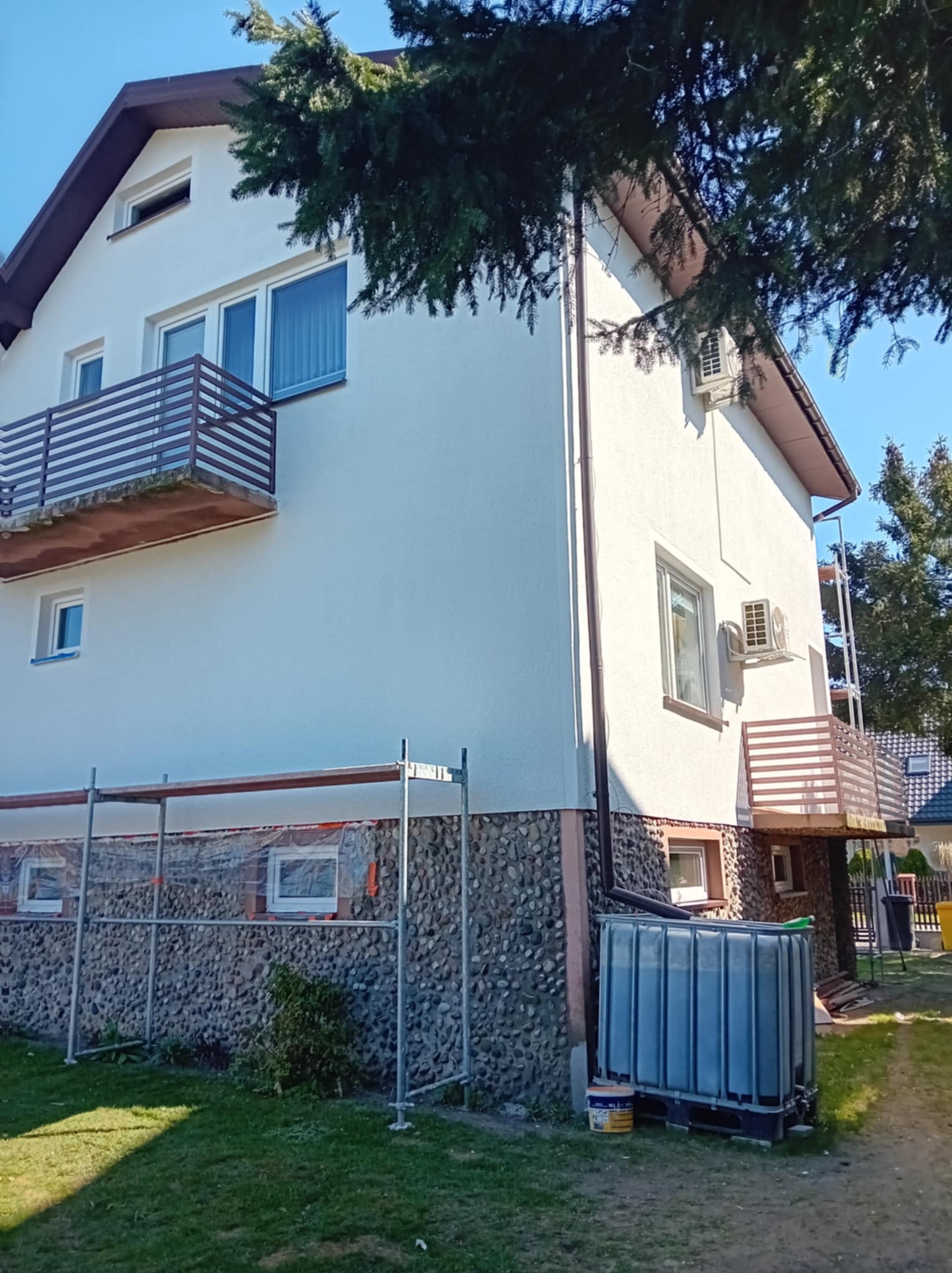 Dwupiętrowy dom z białymi ścianami i kamienną podstawą, częściowo zakryty rusztowaniem. Widoczne balkony z brązowymi barierkami i klimatyzatory na elewacji.