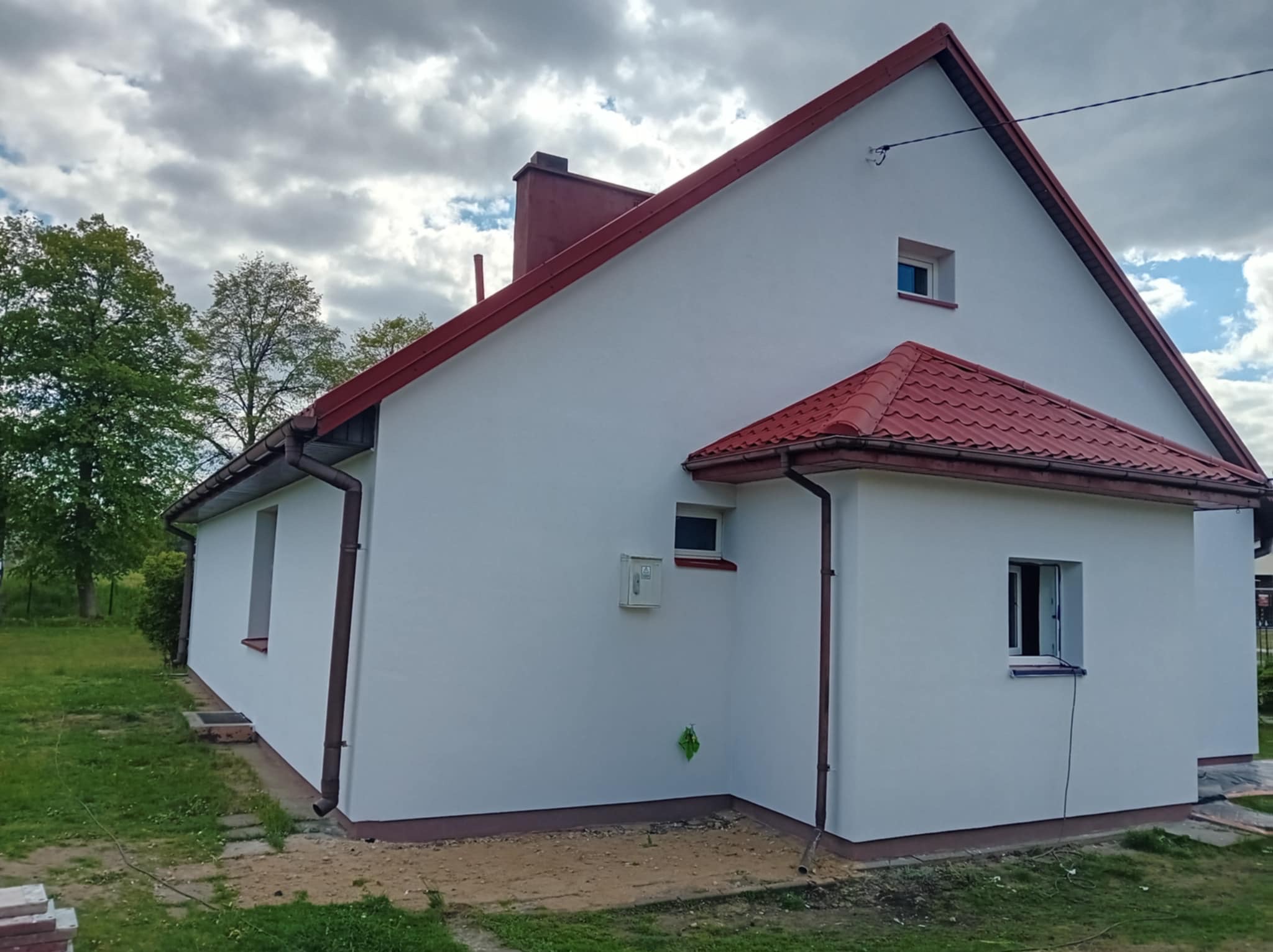 Biały dom jednorodzinny z czerwoną dachówką i rynnami, po renowacji elewacji, widok z boku na tle pochmurnego nieba i zieleni.