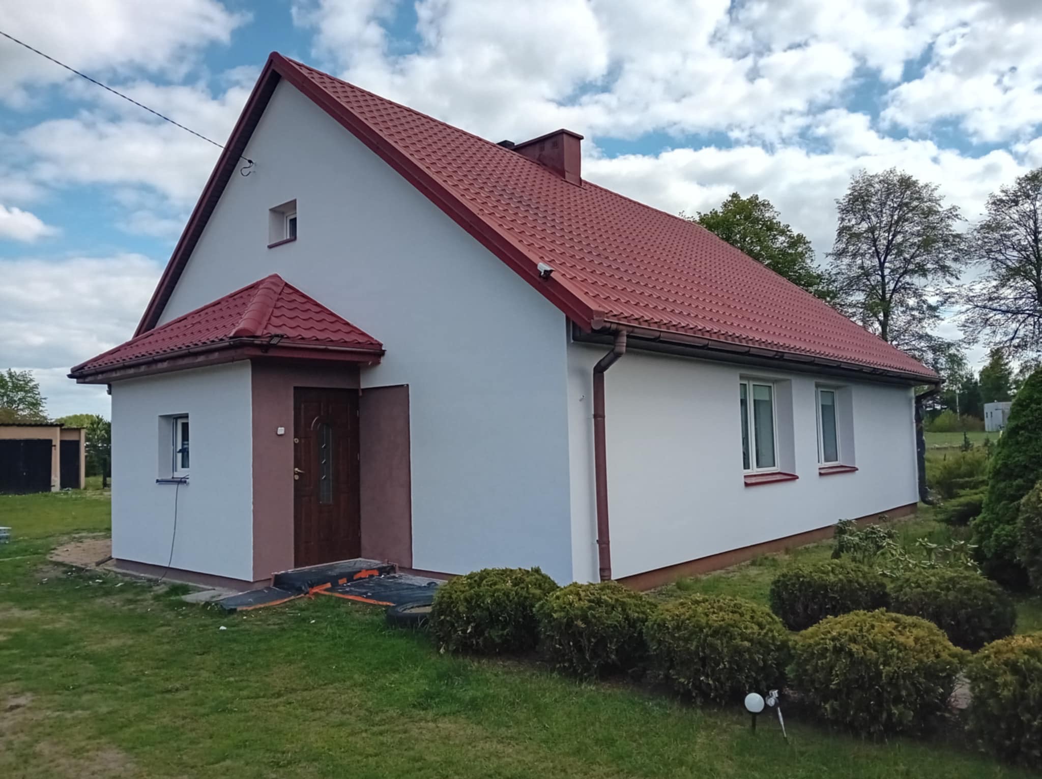 Dom jednorodzinny po remoncie elewacji, z nową, czerwoną dachówką i białymi ścianami. Widoczne elementy wykończeniowe w kolorze brązowym, dopasowane do stolarki okiennej i drzwiowej.