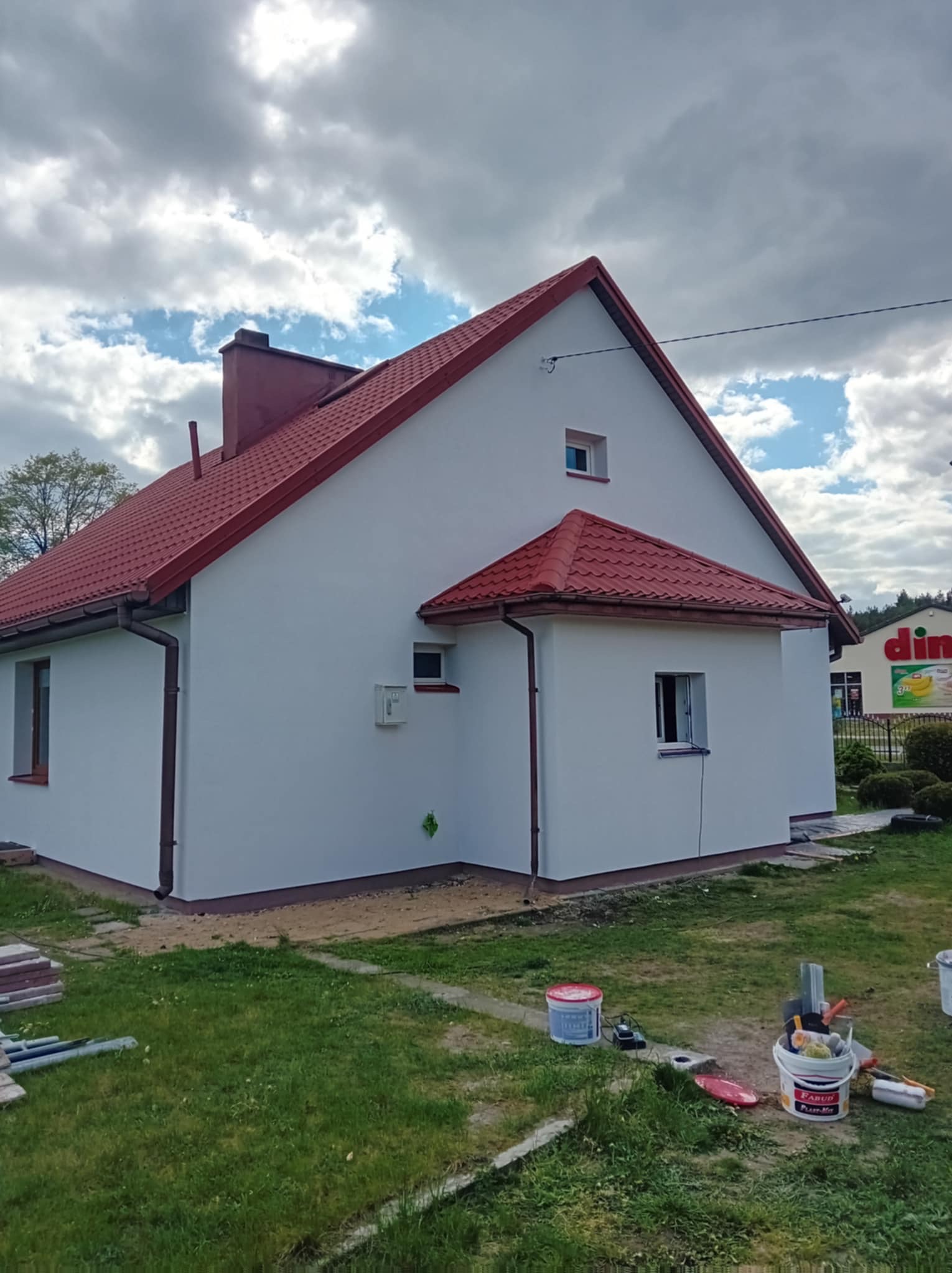 Boczny widok odnowionego domu z czerwoną dachówką i białą elewacją. Na trawie widoczne puszki z farbą i narzędzia malarskie, sugerujące świeżo zakończone prace.
