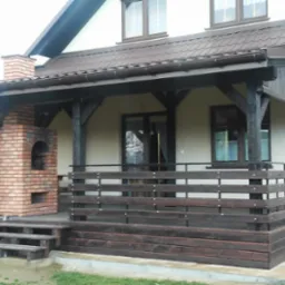 Zadaszony taras z drewnianą balustradą i ceglanym grillem, z trzema schodkami wejściowymi, przylegający do ściany domu z brązowym dachem.