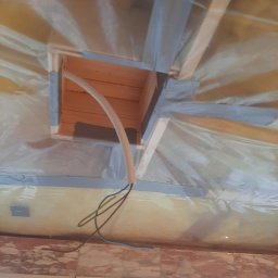 Blue King Sp. Z o.o. - Wnętrze w trakcie budowy, widoczna drewniana konstrukcja stropu z izolacją, peszel z przewodami wychodzący z otworu w suficie, fragment okna w tle.