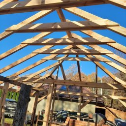 Blue King Sp. Z o.o. - Konstrukcja drewniana z belek, wznoszona na fundamencie z czerwonej cegły, widok od dołu na błękitne niebo, w tle częściowo widoczny dom i drzewa.