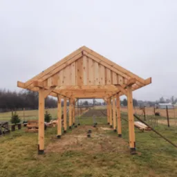 Konstrukcja z surowego drewna w trakcie budowy, prawdopodobnie zadaszenie tarasu lub pergola, widok zewnętrzny na tle zaoranego pola i zabudowań.