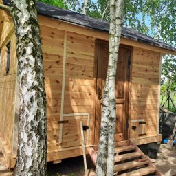 Blue King Sp. Z o.o. - Niewielki drewniany budynek z ciemnym dachem, otoczony brzozami, z widocznymi schodkami wejściowymi i elementami konstrukcyjnymi. Na ścianie widoczny biały kabel zwisający wzdłuż ściany.