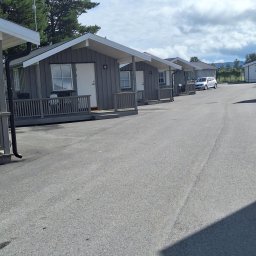 Blue King Sp. Z o.o. - Rząd niewielkich, szarych domków z drewnianą elewacją i białymi ramami okiennymi, oznaczonych numerami, z małym tarasem przed wejściem, położonych wzdłuż asfaltowej drogi.