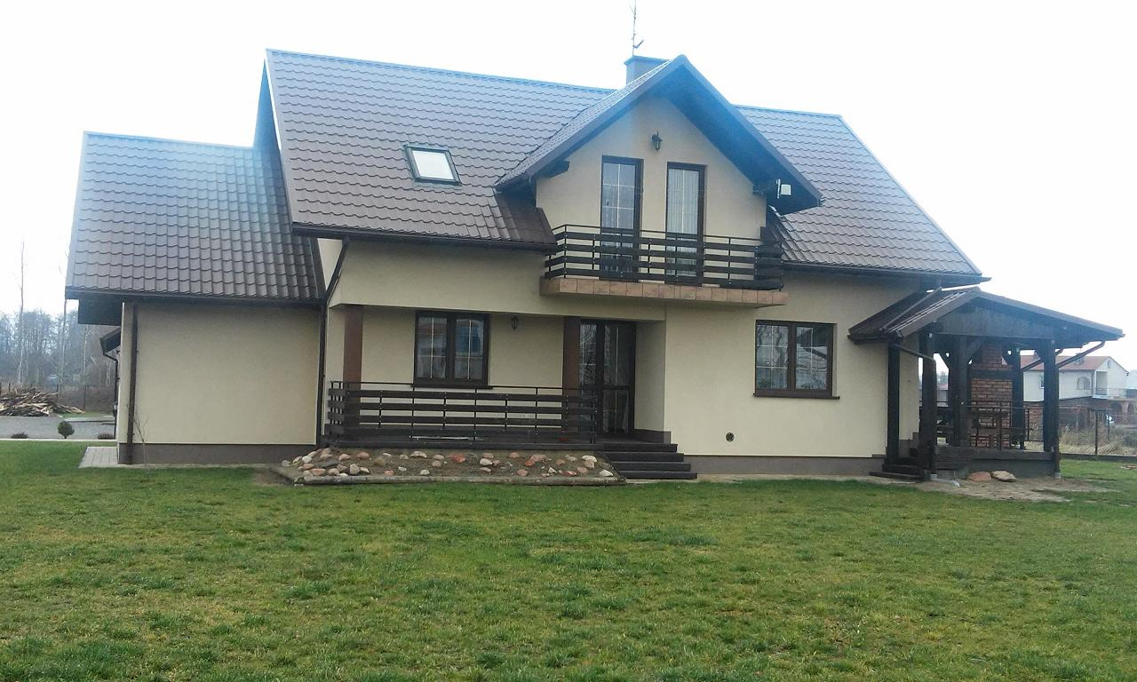 Gotowy dom jednorodzinny z balkonem, brązowym dachem i zadaszonym miejscem do grillowania na zewnątrz, otoczony zielonym trawnikiem.