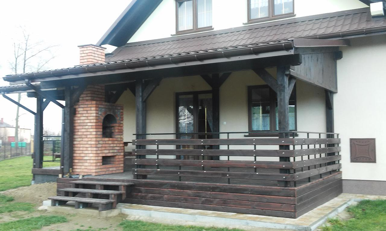 Zadaszony taras z drewnianą balustradą i ceglanym grillem, z trzema schodkami wejściowymi, przylegający do ściany domu z brązowym dachem.