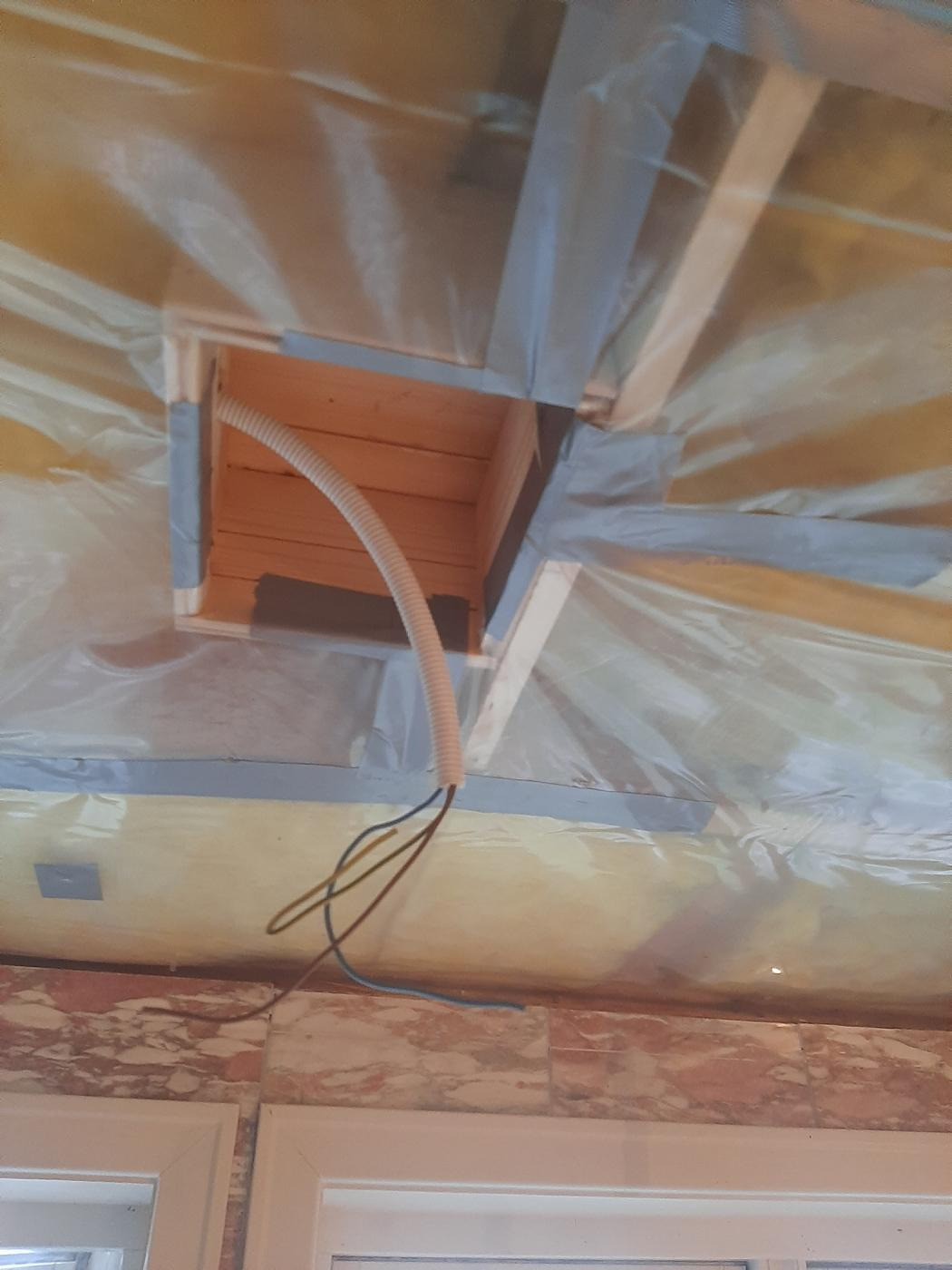 Wnętrze w trakcie budowy, widoczna drewniana konstrukcja stropu z izolacją, peszel z przewodami wychodzący z otworu w suficie, fragment okna w tle.