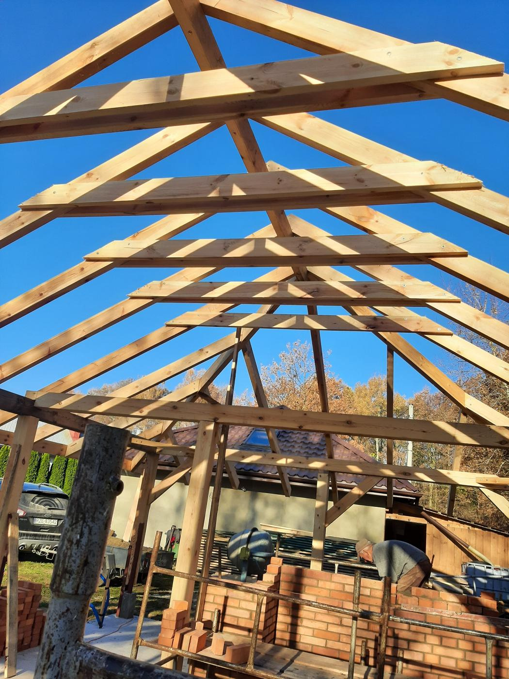 Konstrukcja drewniana z belek, wznoszona na fundamencie z czerwonej cegły, widok od dołu na błękitne niebo, w tle częściowo widoczny dom i drzewa.