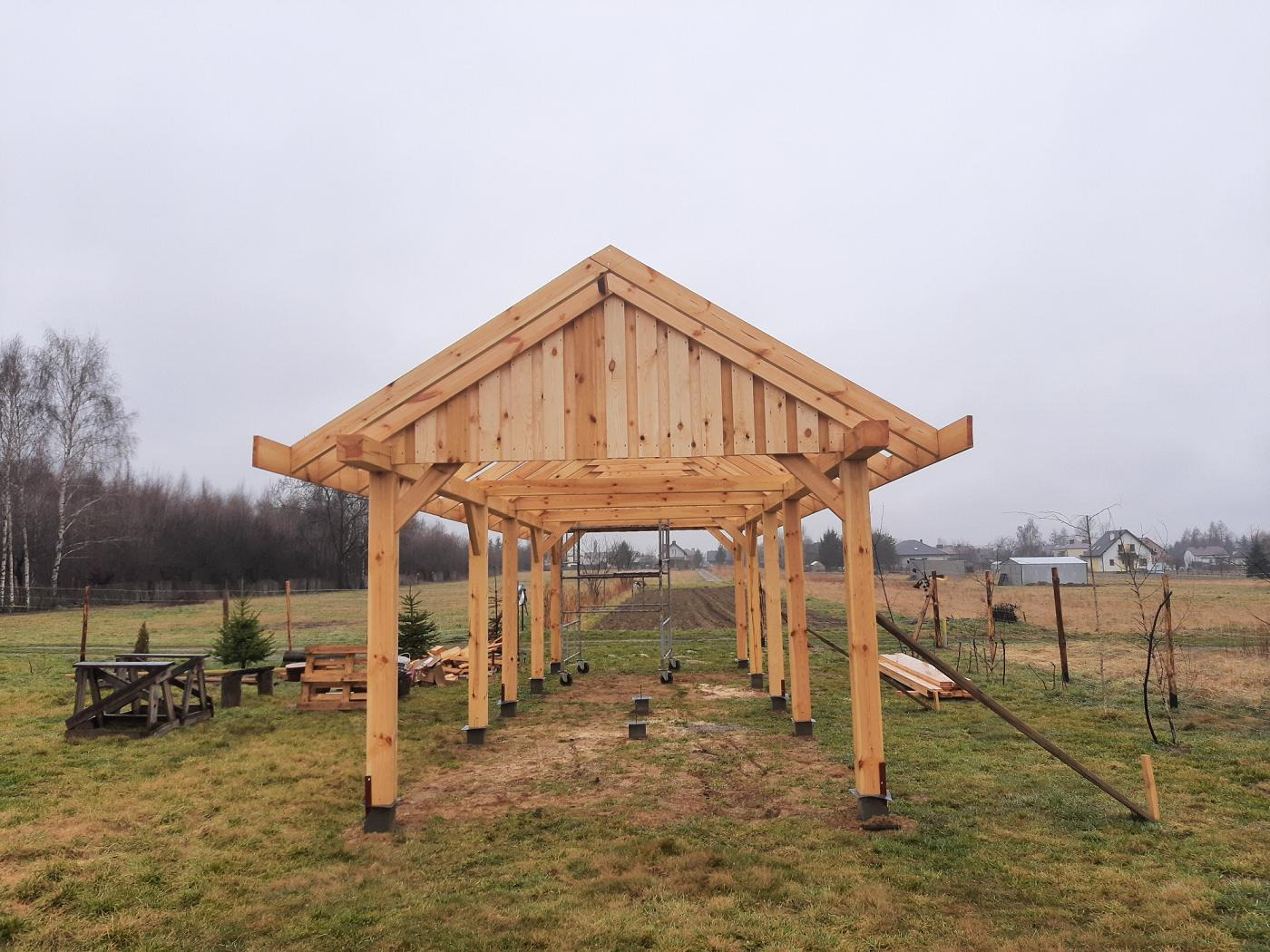 Konstrukcja z surowego drewna w trakcie budowy, prawdopodobnie zadaszenie tarasu lub pergola, widok zewnętrzny na tle zaoranego pola i zabudowań.