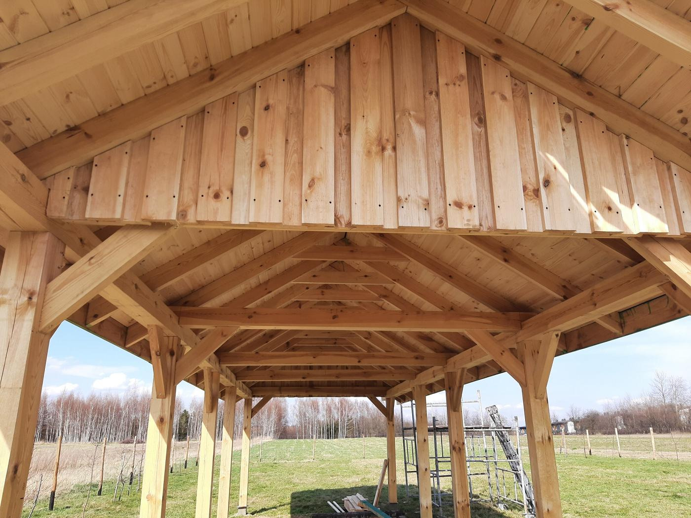 Konstrukcja altany z surowego drewna, widok od wewnątrz, z widocznym fragmentem krajobrazu w tle i elementami rusztowania.