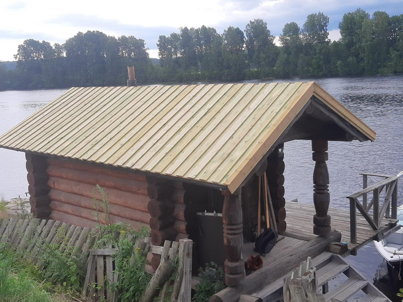 Mały drewniany domek z dachem z desek, stojący na drewnianym pomoście nad wodą; w tle widać rzekę i drzewa.