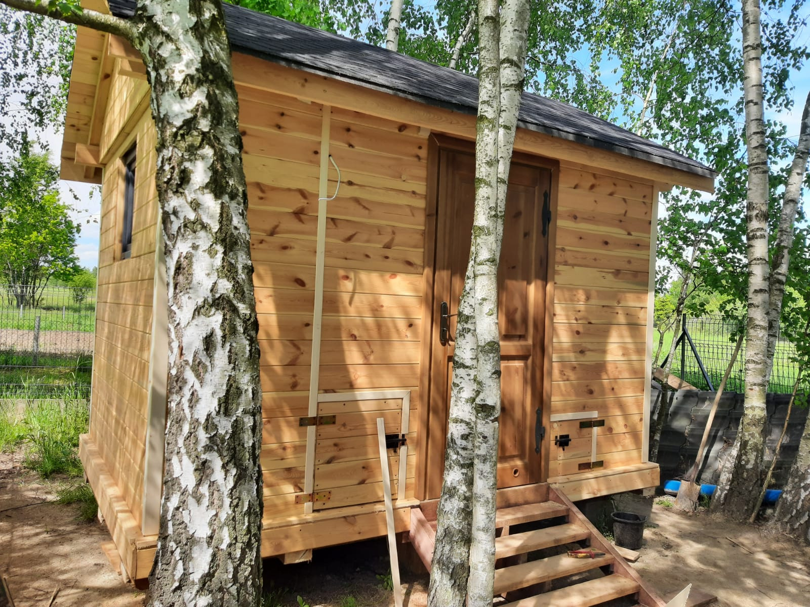 Niewielki drewniany budynek z ciemnym dachem, otoczony brzozami, z widocznymi schodkami wejściowymi i elementami konstrukcyjnymi. Na ścianie widoczny biały kabel zwisający wzdłuż ściany.