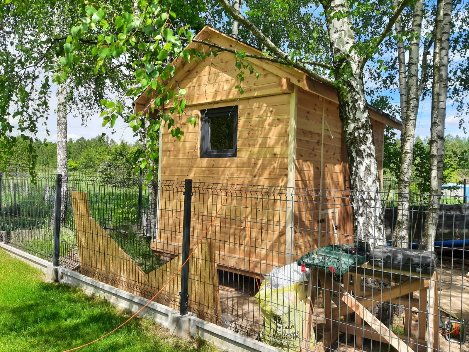 Drewniany kurnik z czarnym oknem, częściowo ukryty za brzozą, otoczony siatką ogrodzeniową. Widoczna wełna mineralna i narzędzia na zewnątrz.