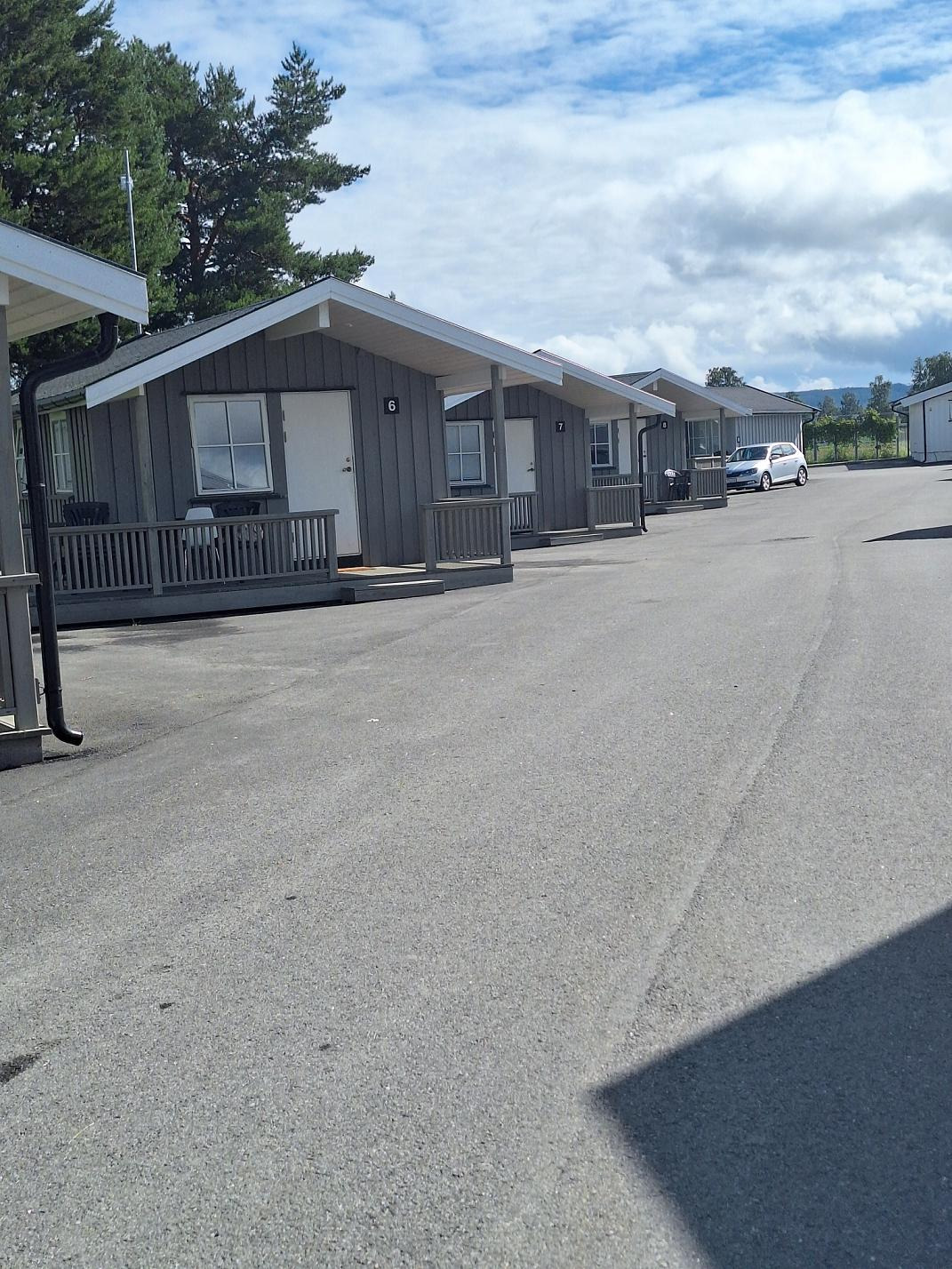 Rząd niewielkich, szarych domków z drewnianą elewacją i białymi ramami okiennymi, oznaczonych numerami, z małym tarasem przed wejściem, położonych wzdłuż asfaltowej drogi.