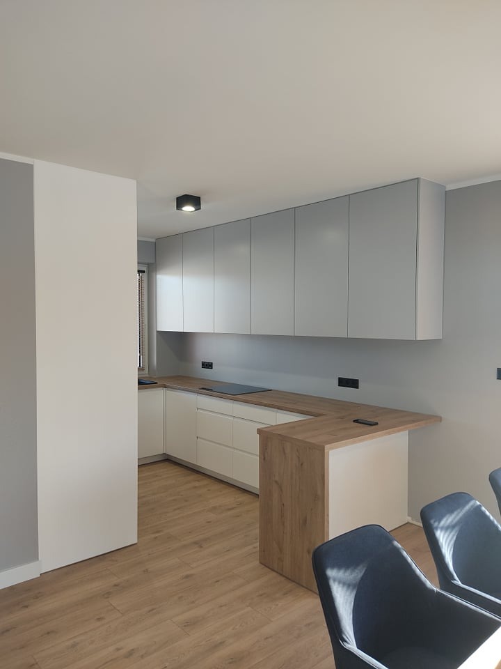 Nowoczesna kuchnia z białymi szafkami dolnymi i szarymi górnymi, drewniany blat z barkiem, panele podłogowe i fragment krzeseł w tle.