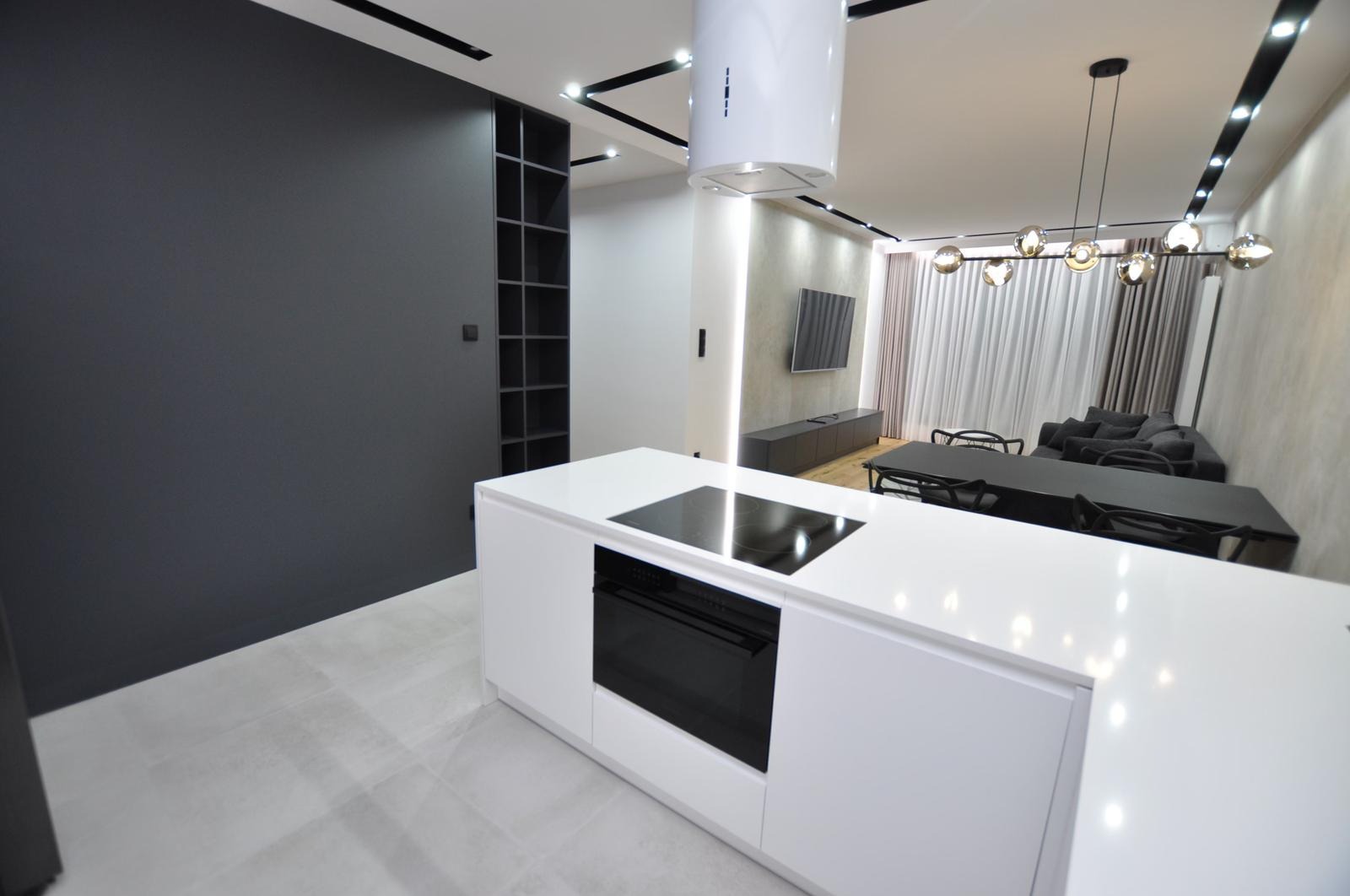 Nowoczesne wnętrze: biała wyspa kuchenna z czarną płytą indukcyjną i piekarnikiem, szara ściana z czarnym regałem, oświetlenie punktowe i wisząca lampa nad stołem jadalnym, salon w tle z szarą sofą...