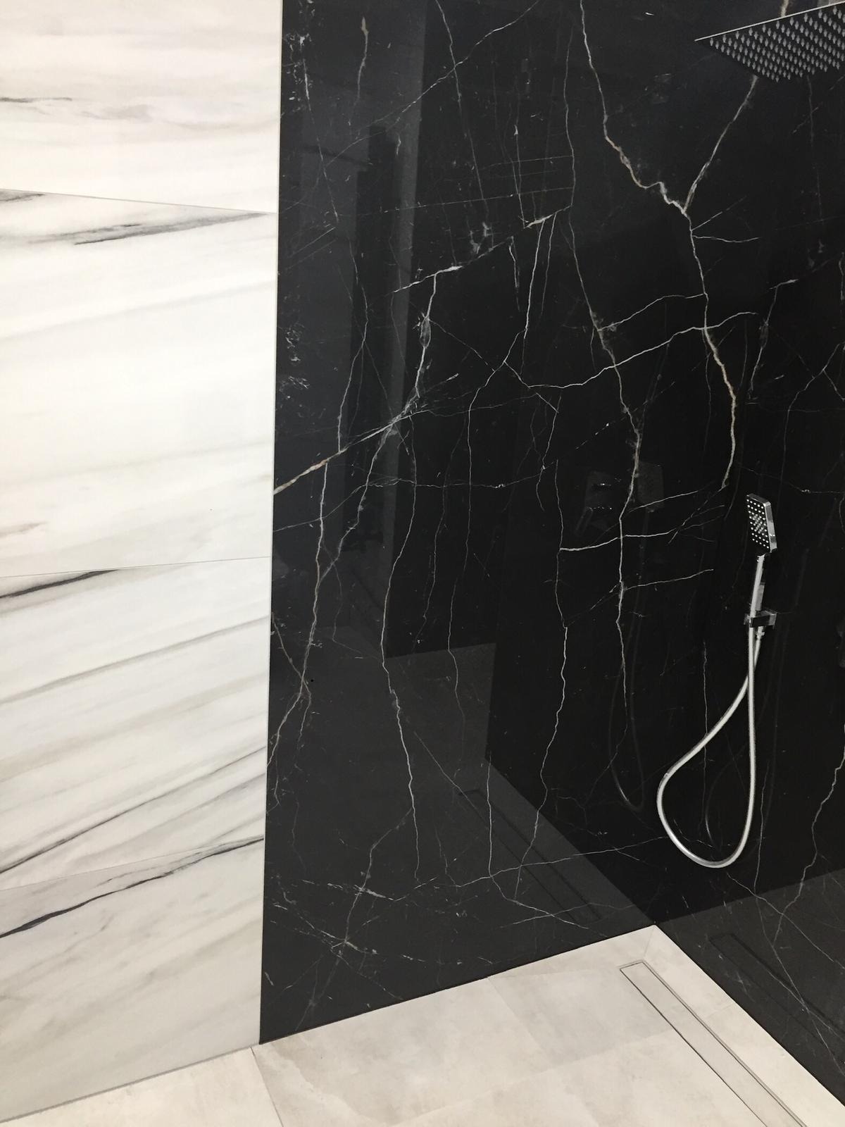 Ściana wyłożona czarnymi i białymi płytami imitującymi marmur, z widocznym odpływem liniowym i słuchawką prysznicową na tle czarnej płyty.