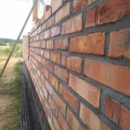 Świeżo wymurowana ściana z czerwonej cegły z widoczną izolacją fundamentu i poziomowaniem na budowie.