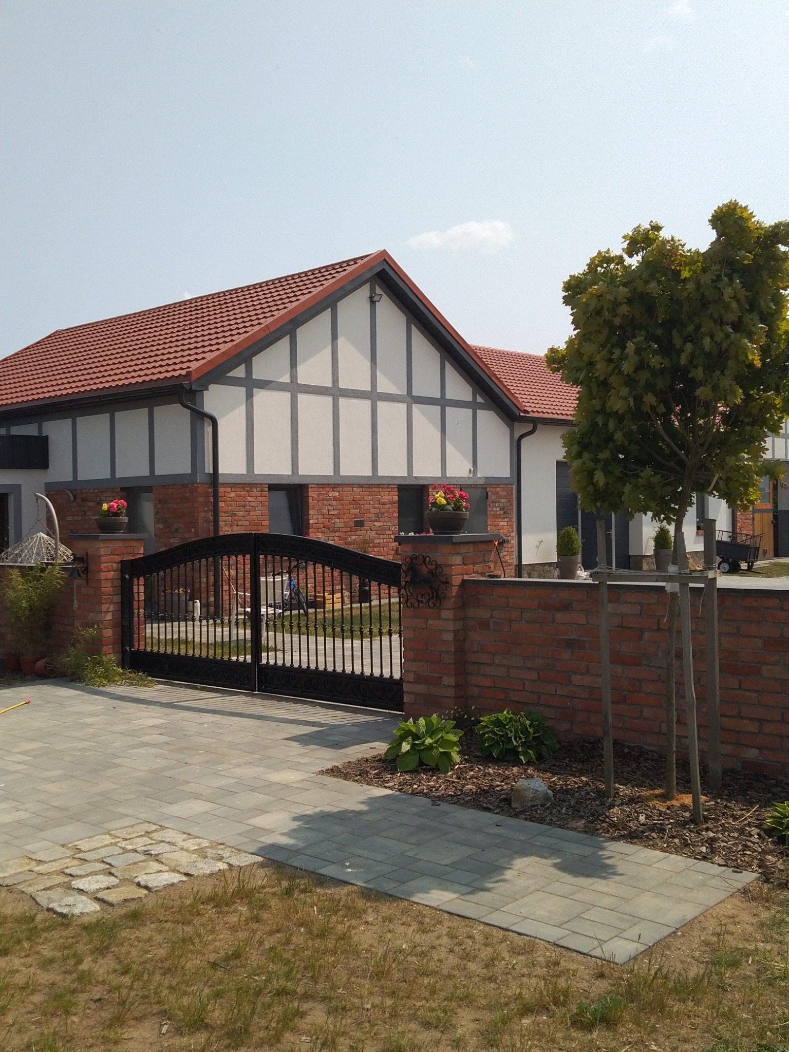 Dom jednorodzinny z czerwoną dachówką, białą elewacją z szarymi elementami dekoracyjnymi, ogrodzony ceglanym murem z czarną bramą, rabaty z roślinami i kostka brukowa na podjeździe.