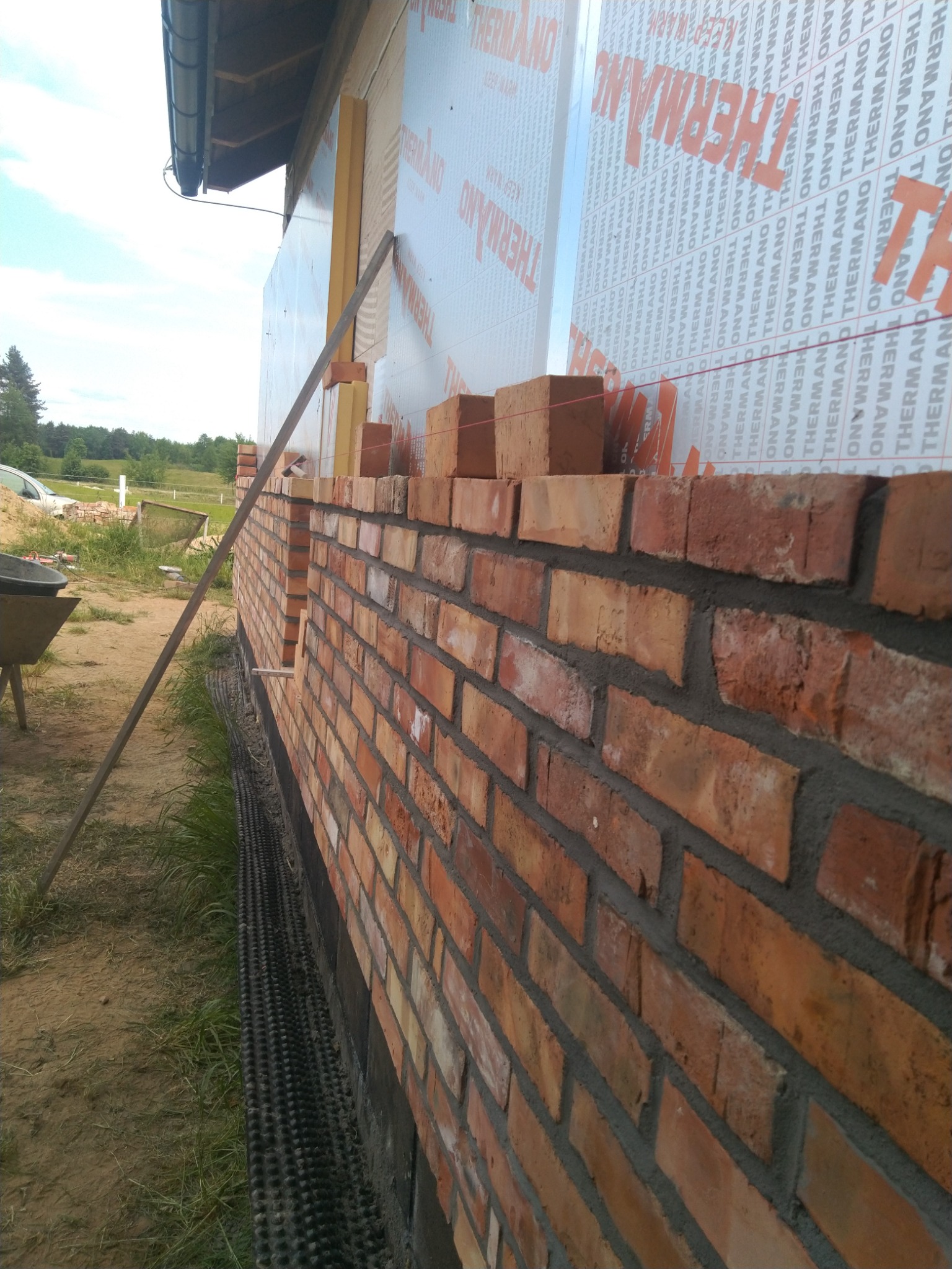 Wznoszenie ściany z czerwonej cegły na zewnątrz budynku, widoczne ocieplenie ściany z napisem THERMANO, poziomica oparta o ścianę, sznurek murarski wyznaczający linię, teren budowy w tle.