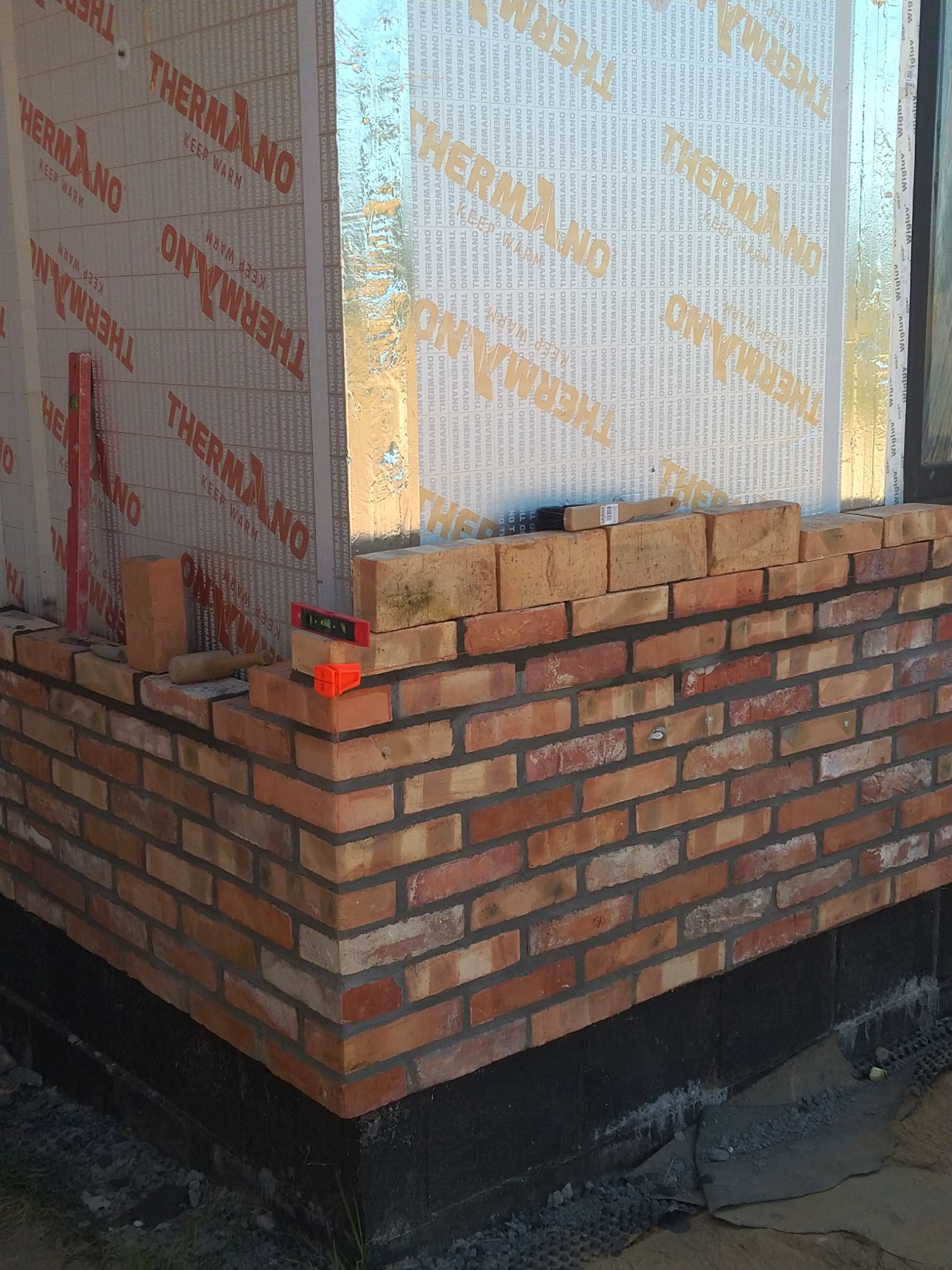 Murowanie ściany z cegły klinkierowej na ocieplonym fundamencie, widoczne poziomice i narzędzia murarskie.