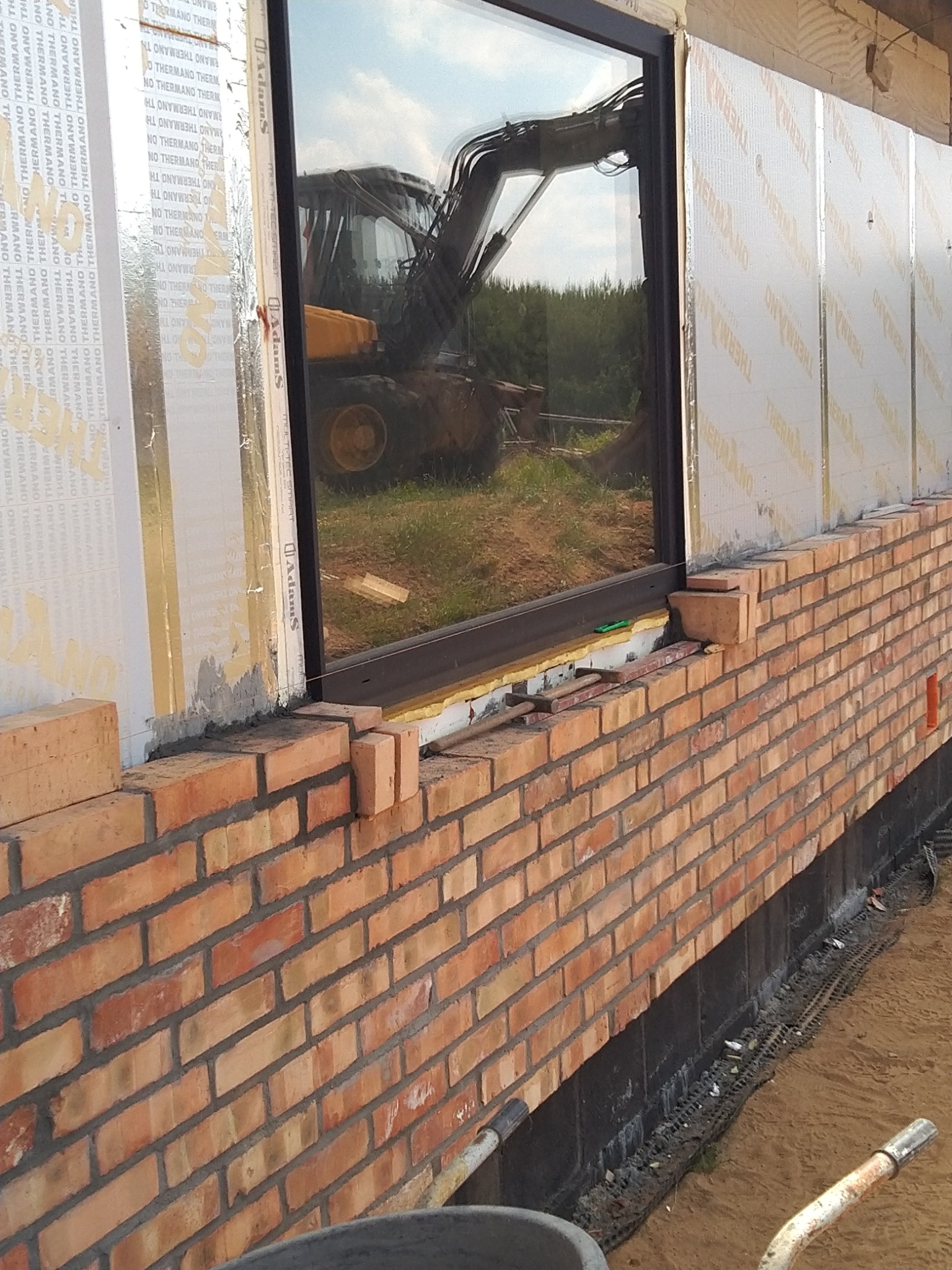 Murowana ściana z cegły klinkierowej w trakcie budowy, widoczna izolacja termiczna i odbicie koparki w szybie okiennej.