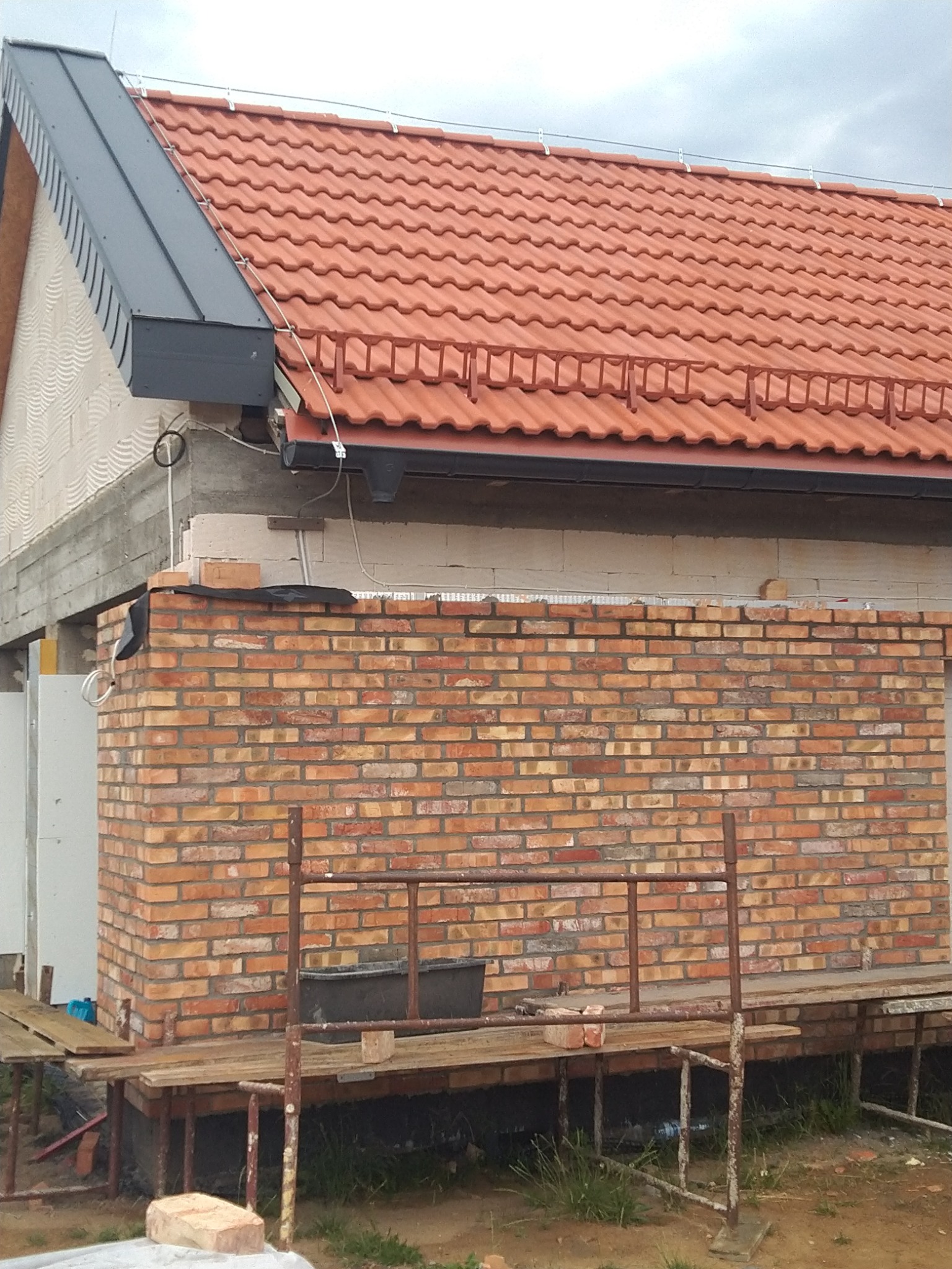 Wznoszenie ściany z cegły klinkierowej na budowie domu jednorodzinnego, widoczne rusztowanie i fragment dachu z dachówki ceramicznej.