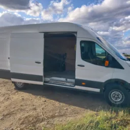 Biały bus z otwartymi drzwiami bocznymi, zaparkowany na polnej drodze w Warszawie, z widocznym wnętrzem przedziału ładunkowego.