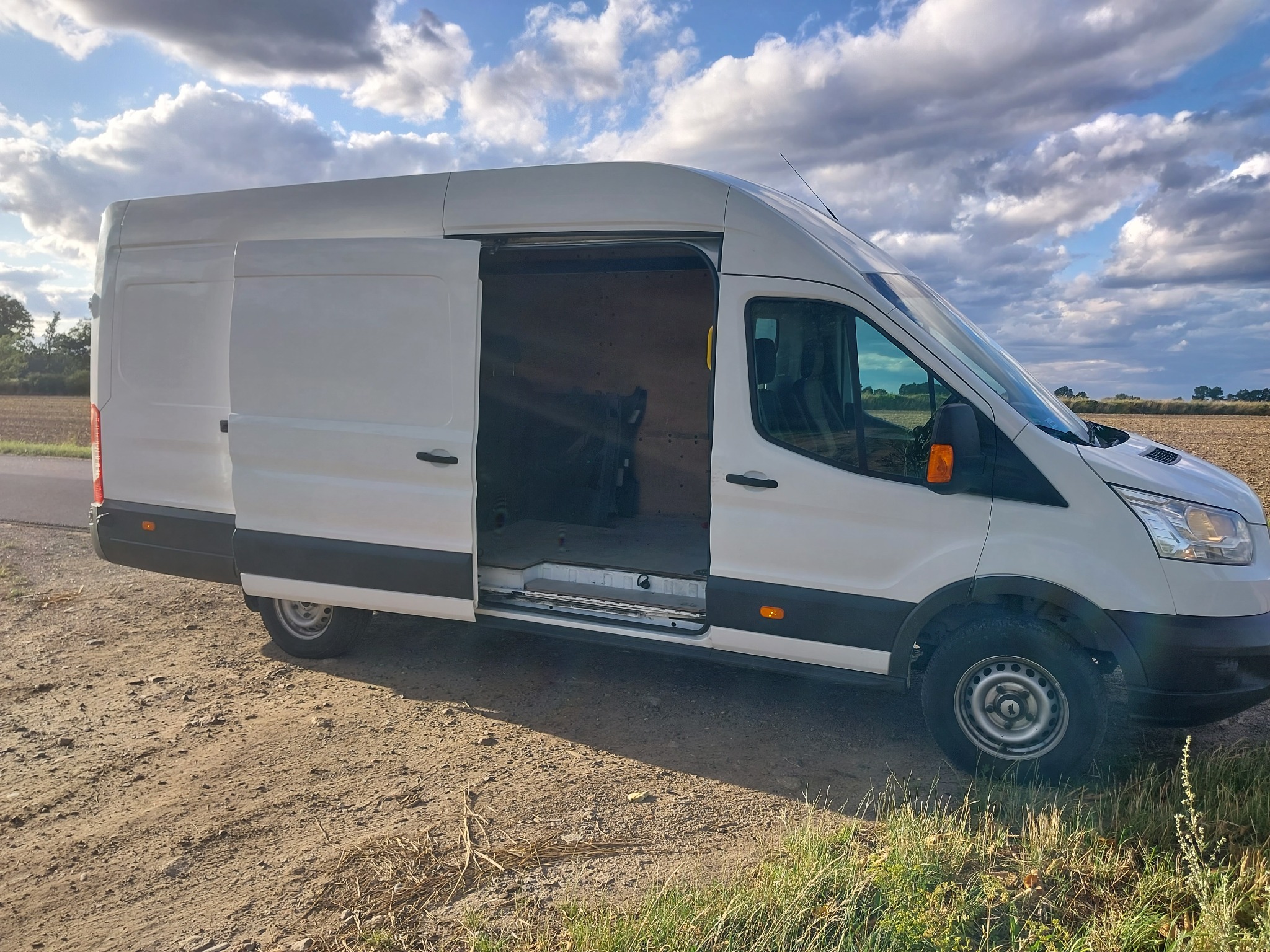 Biały bus z otwartymi drzwiami bocznymi, zaparkowany na polnej drodze w Warszawie, z widocznym wnętrzem przedziału ładunkowego.