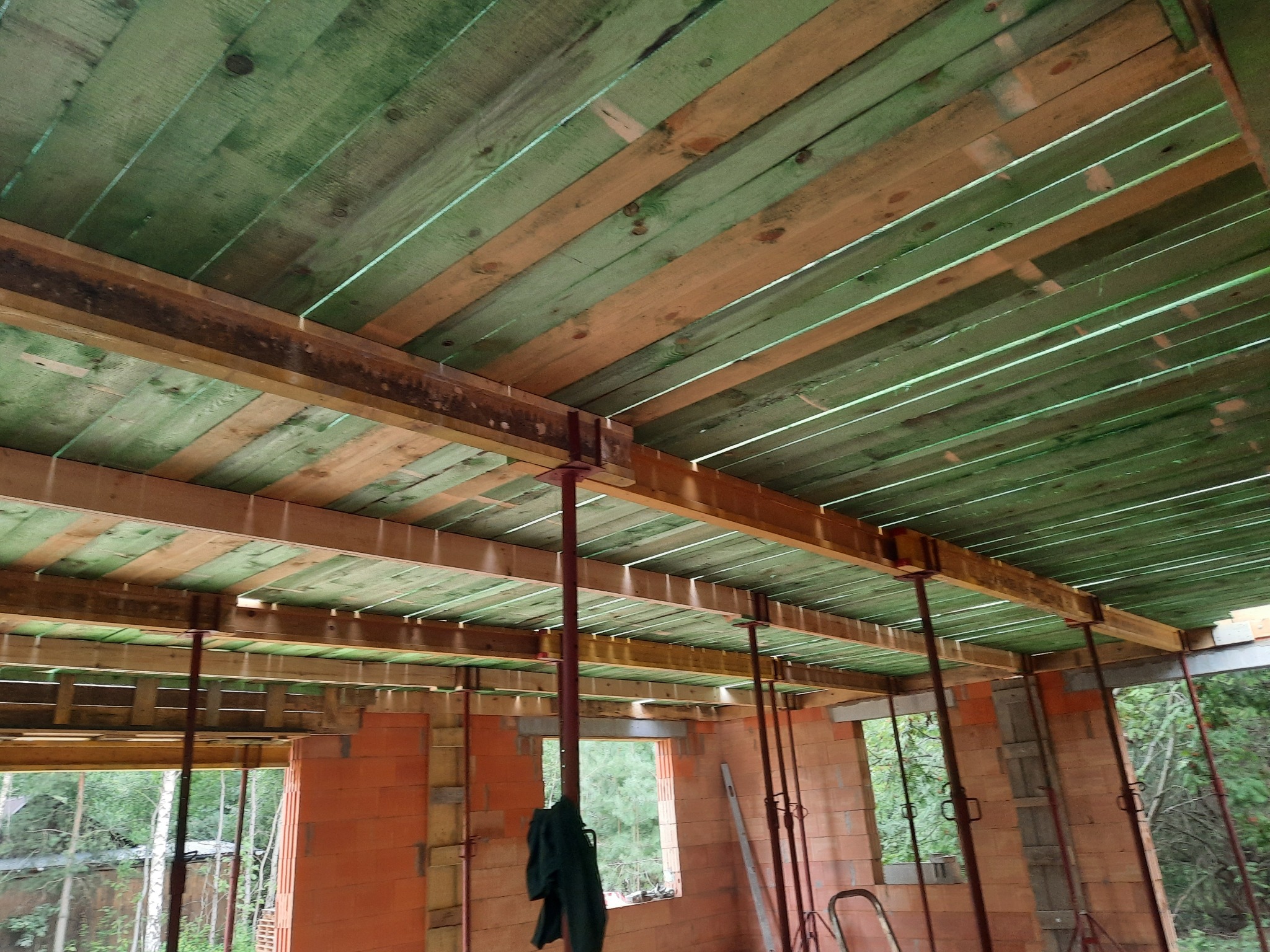 Wnętrze wznoszonego domu z czerwonej cegły z widocznym szalunkiem stropu, wspartym na metalowych stemplach, z zielonymi deskami wypełniającymi przestrzeń między belkami.