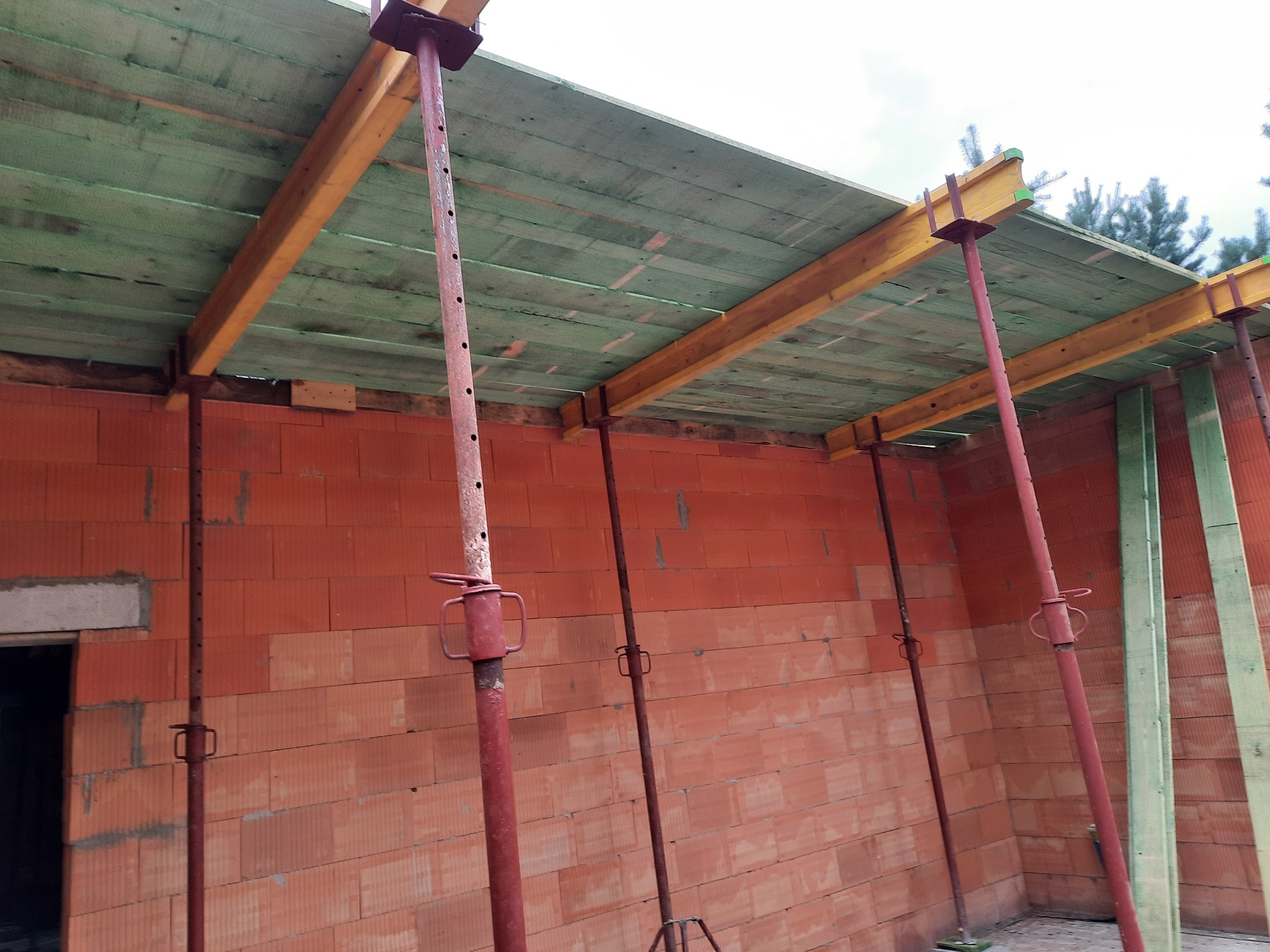 Wznoszenie muru z czerwonej cegły z widocznym szalunkiem stropowym podpartym metalowymi stemplami budowlanymi. Widoczny otwór drzwiowy w ścianie.