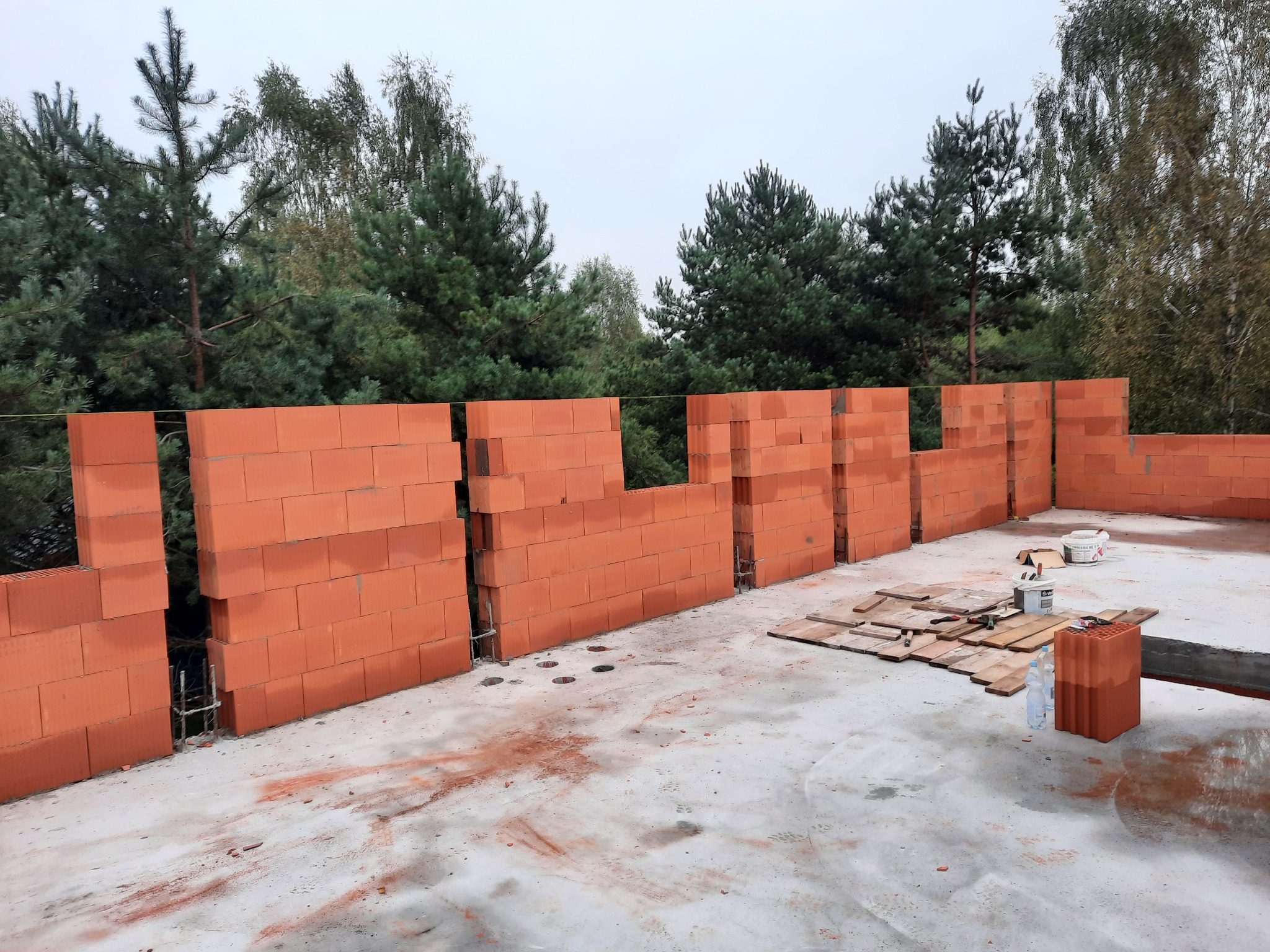 Wznoszenie ścian parteru z czerwonej cegły na fundamencie, widoczne zbrojenia pionowe, w tle drzewa iglaste i liściaste.