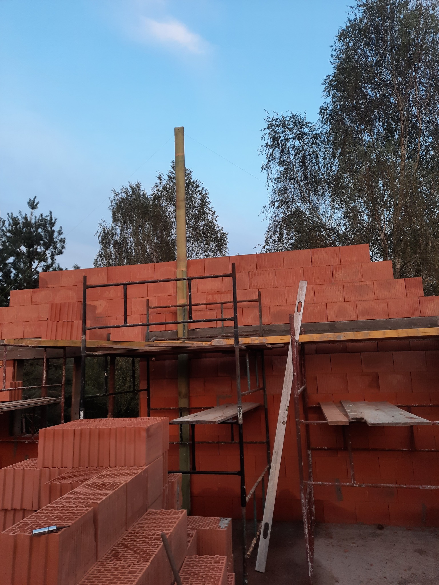 Wznoszenie muru z czerwonych bloczków ceramicznych na rusztowaniu, widoczna poziomica i zapas materiału budowlanego, drzewa w tle.